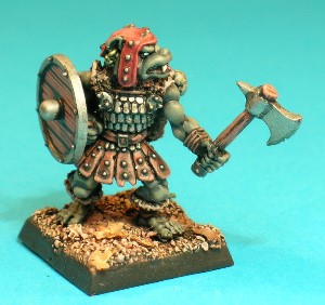 Pose 1, variant A. This Gnoll warrior wears a skirt made of studded leather strips and a scalemail tunic adorned with tatty furs. He carries a sheathed shortsword on his back and is armed with a hand axe in his left hand and a shield in his right. This variant wears a studded leather cap with long earflaps and his axe has a notched single blade and rear spike. He has a dog-like face with a short muzzle, and a part-open mouth revealing upper fangs and a small tongue. He looks to his left and has a fancy earring in each ear.