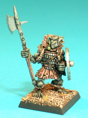 Pose 3, variant A. This Gnoll wears a skirt made of studded leather strips, a tunic of small overlapping steel plates, and a ratty old fur waistcoat. He carries a scabbarded broadsword strapped to his back, he protects himself with a shield on his left arm and holds an upright pole-weapon in his right hand. This variant is bare-headed, revealing a short mohican-style haircut. His polearm is a halberd, with a broad, notched axe-blade on the far side (in relation to the Gnoll) and a short spike on the nearside. The Gnoll has a dog-like face with a short muzzle, and a snarling mouth revealing upper fangs and a set of teeth. His head is tilted slightly back and he looks over his left shoulder. He has an earring with a hanging disc (a dog-tag?) in his right ear and 2 plain rings in his left.